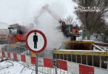 Maszyna budowlana z podnośnikiem pracuje na placu budowy, wydobywając parę z wykopu, obok stoi znak zakazu wstępu.
