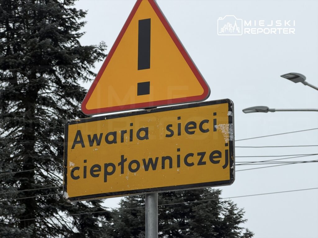Żółty znak ostrzegawczy informujący o awarii sieci ciepłowniczej w otoczeniu drzew i ulicznych latarni.