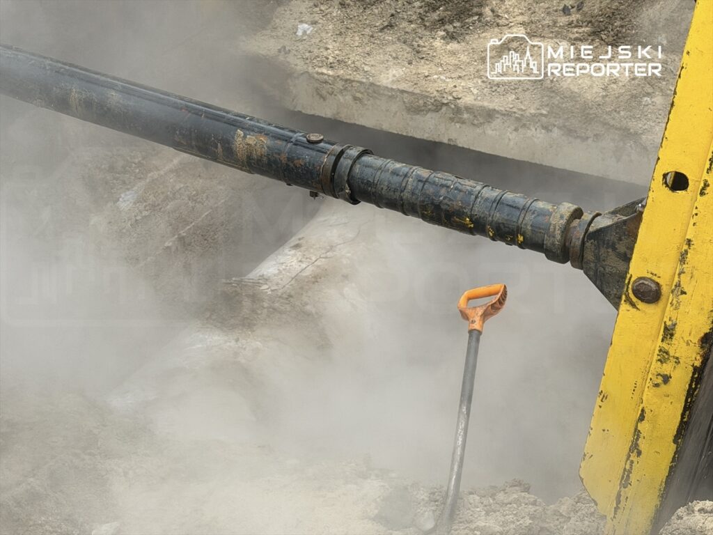 Czarna rura hydrauliczna wydobywająca parę z wykopu budowlanego, obok leży łopata. W powietrzu unosi się pył.