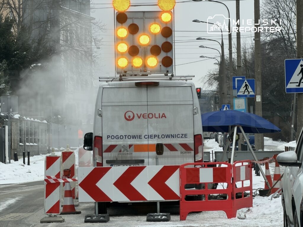 Samochód pogotowia ciepłowniczego z migającymi światłami stoi na zaśnieżonej ulicy, otoczony barierkami.