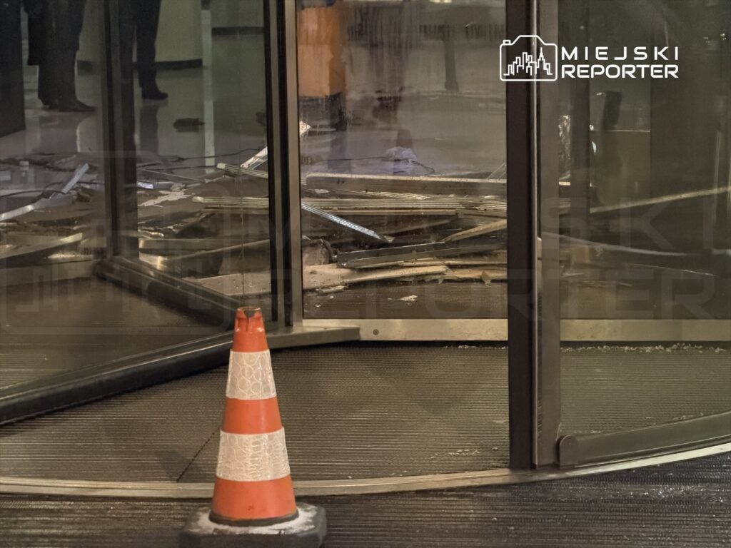 Uszkodzone drzwi obrotowe z rozbitym szkłem w lobby budynku, obok pomarańczowy słupek ostrzegawczy.