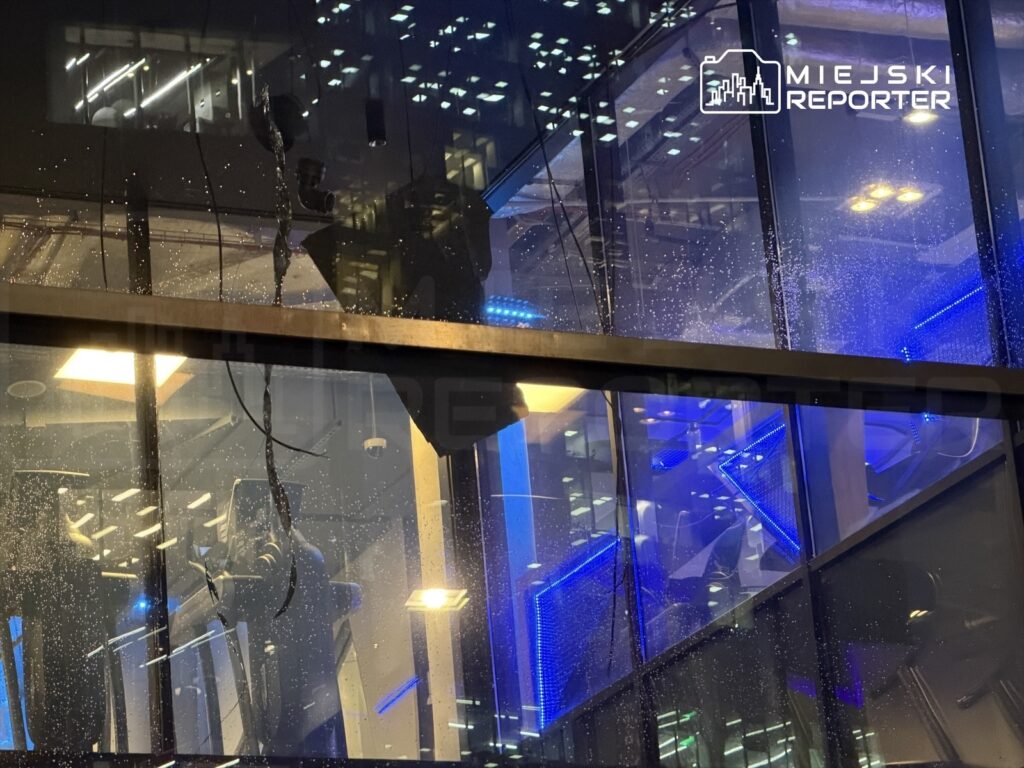 Wnętrze nowoczesnego biurowca z przeszkloną fasadą, oświetlone niebieskimi światłami, z widocznymi kroplami wody na szybie.