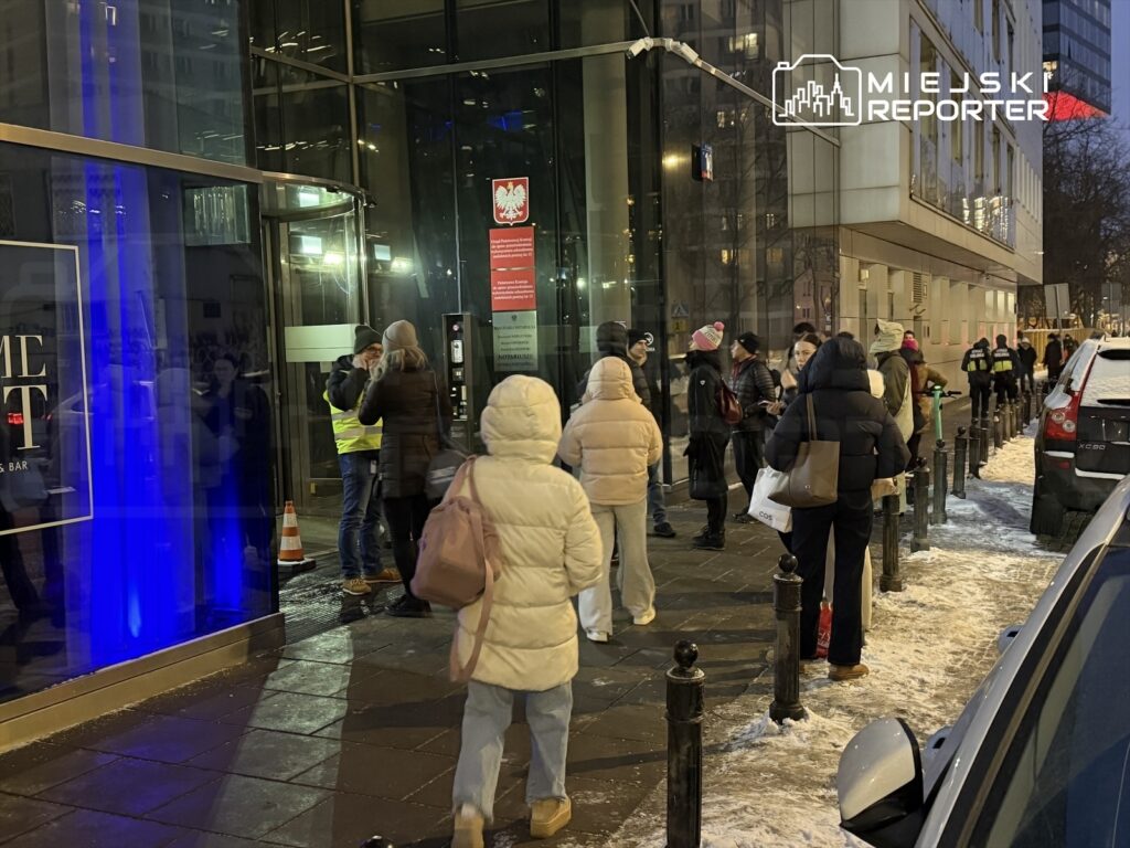 Grupa osób stoi w kolejce przed nowoczesnym budynkiem z przeszkloną fasadą, w nocy, w zimowej scenerii.