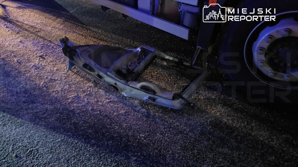 Uszkodzona rama pojazdu leży na jezdni obok dużego samochodu dostawczego, oświetlona niebieskim światłem.