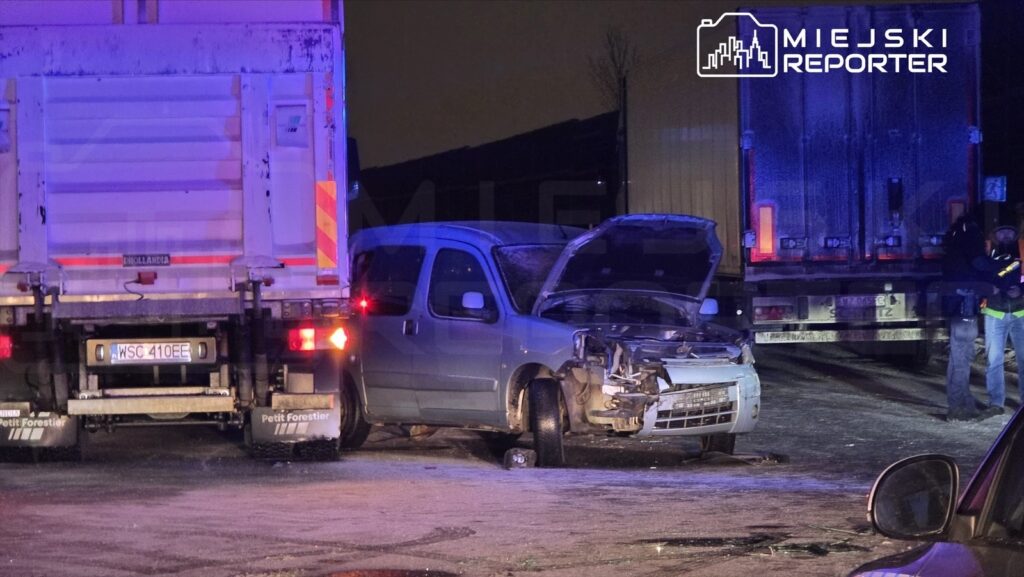Zniszczony samochód dostawczy z uniesioną maską stoi obok ciężarówki na oświetlonym parkingu nocą. Obok grupka osób w kamizelkach odblaskowych.