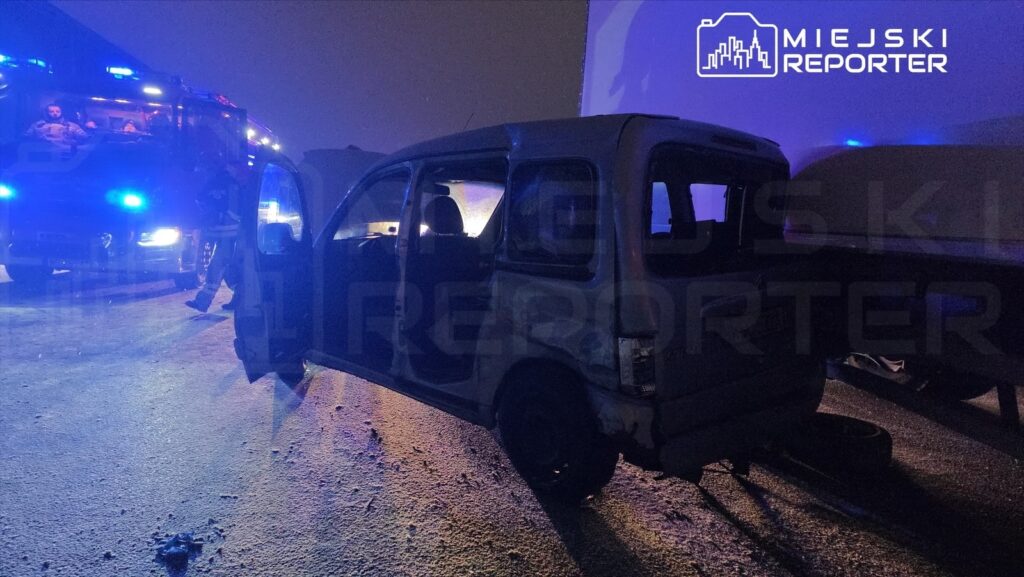 Samochód osobowy z otwartymi drzwiami stoi na zaśnieżonej drodze, obok niego widać wóz strażacki z niebieskimi światłami.