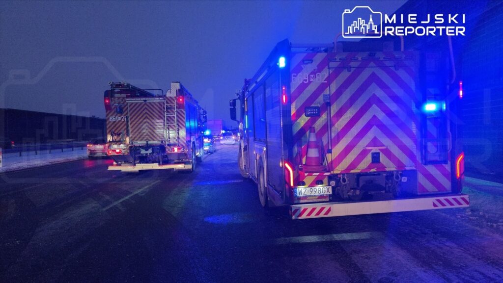 Na drodze w nocy stoją dwa wozy strażackie z włączonymi sygnalizacjami świetlnymi.