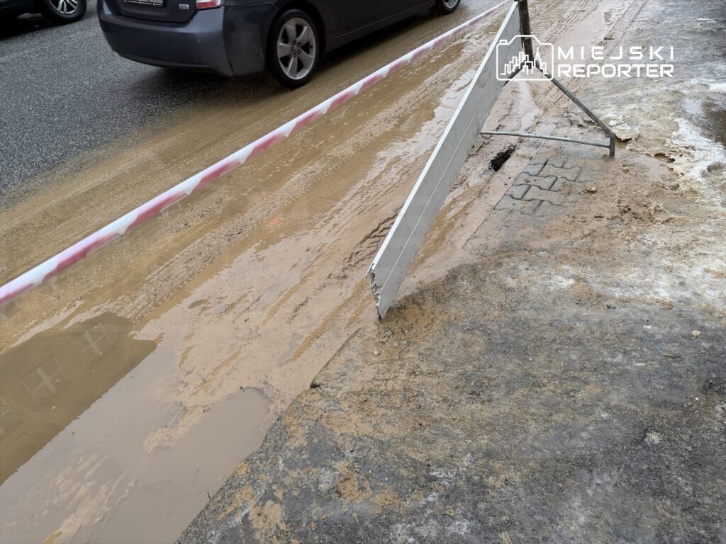 Na drodze widoczna jest kałuża błota oraz barierka ostrzegawcza. W tle przejeżdżające samochody.