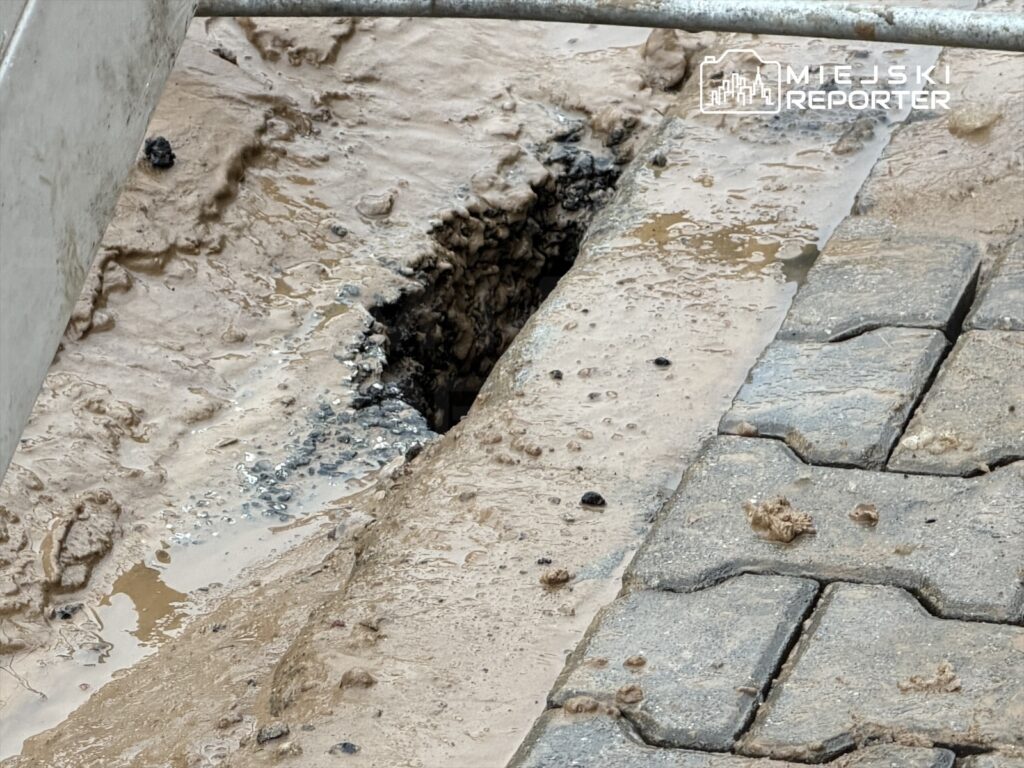 Zniszczona kostka brukowa z widoczną dziurą, wypełnioną wodą i błotem, w miejskim otoczeniu.