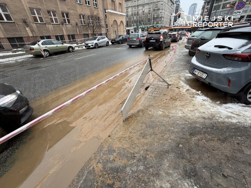 Na jezdni widoczna plama błota obok zaparkowanych samochodów, z barierką ostrzegawczą na chodniku.