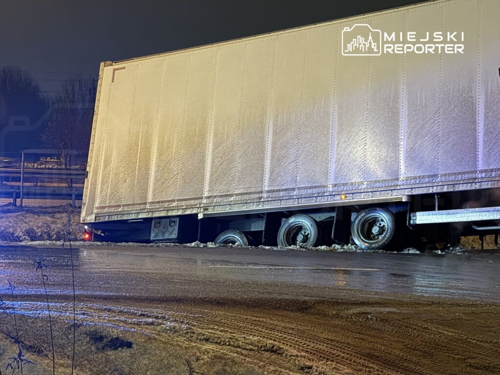 Ciężarówka z naczepą utknęła w błocie na drodze, obok świecących świateł policyjnych w nocy.