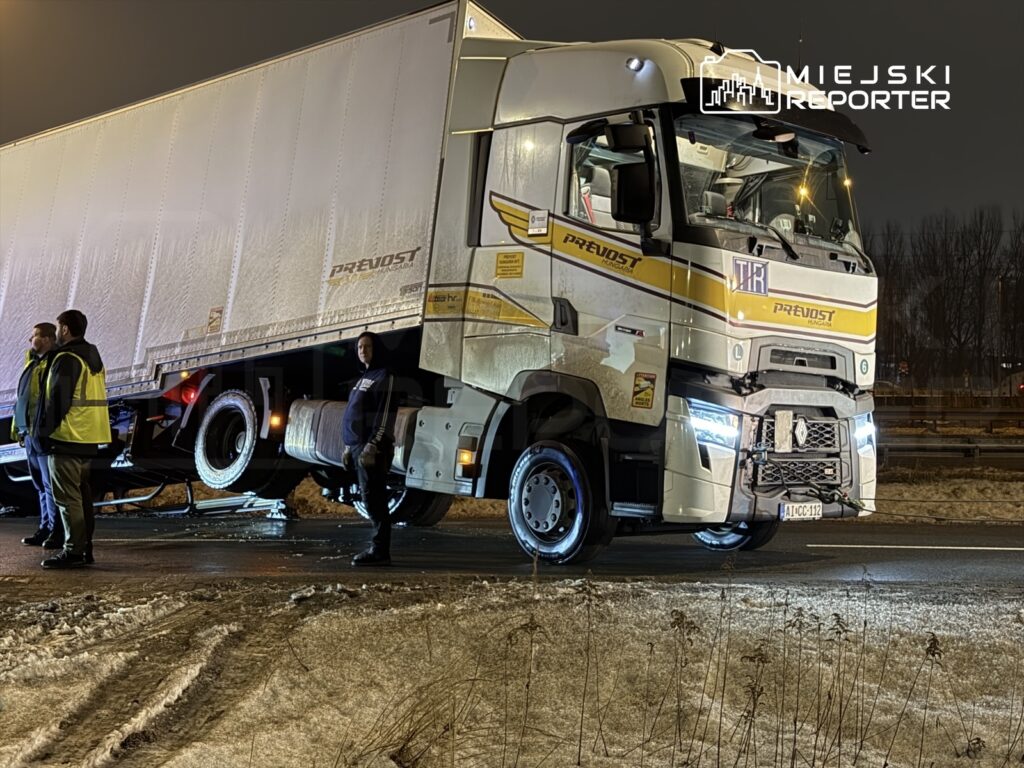 Trzech mężczyzn w odblaskowych kamizelkach stoi obok unieruchomionego ciężarowego pojazdu na nocnej drodze.