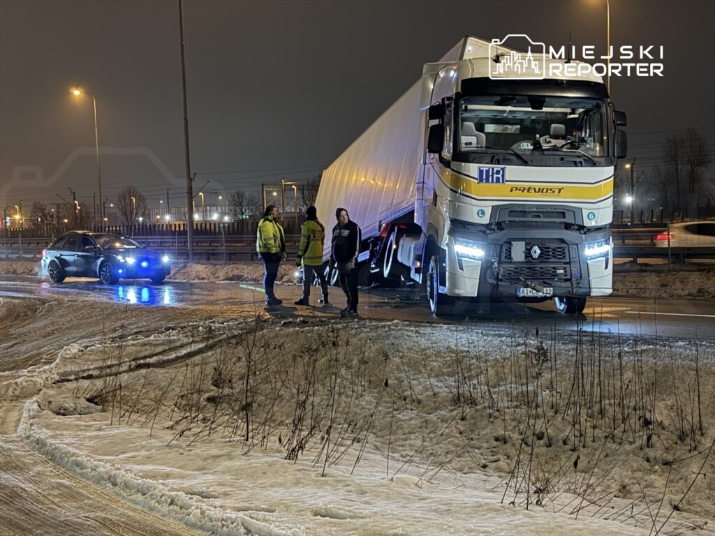 Czterech mężczyzn w odblaskowych kamizelkach rozmawia obok ciężarówki na poboczu drogi w nocy.