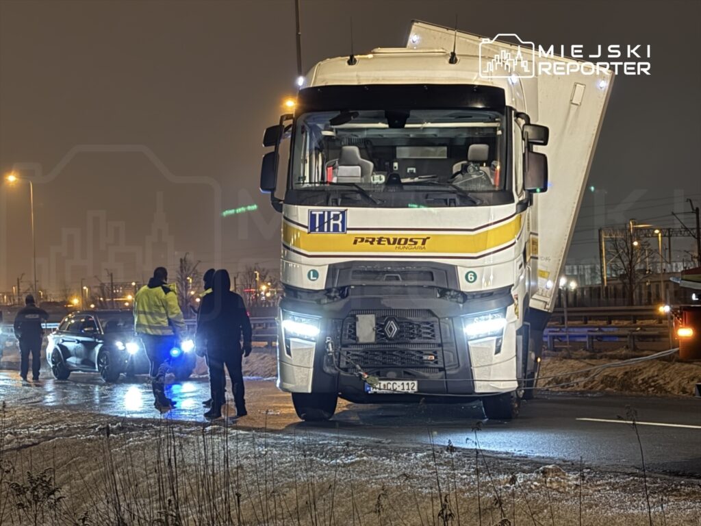 Grupa osób w odblaskowych kamizelkach rozmawia przy zatrzymanym ciężarowym pojeździe na drodze nocą.