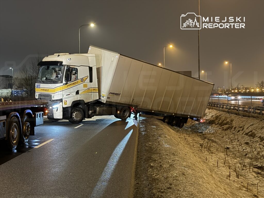 Ciężarówka z naczepą w rowie przy drodze, kierowca próbuje ją wyciągnąć w nocy. W tle widać światła innych pojazdów.