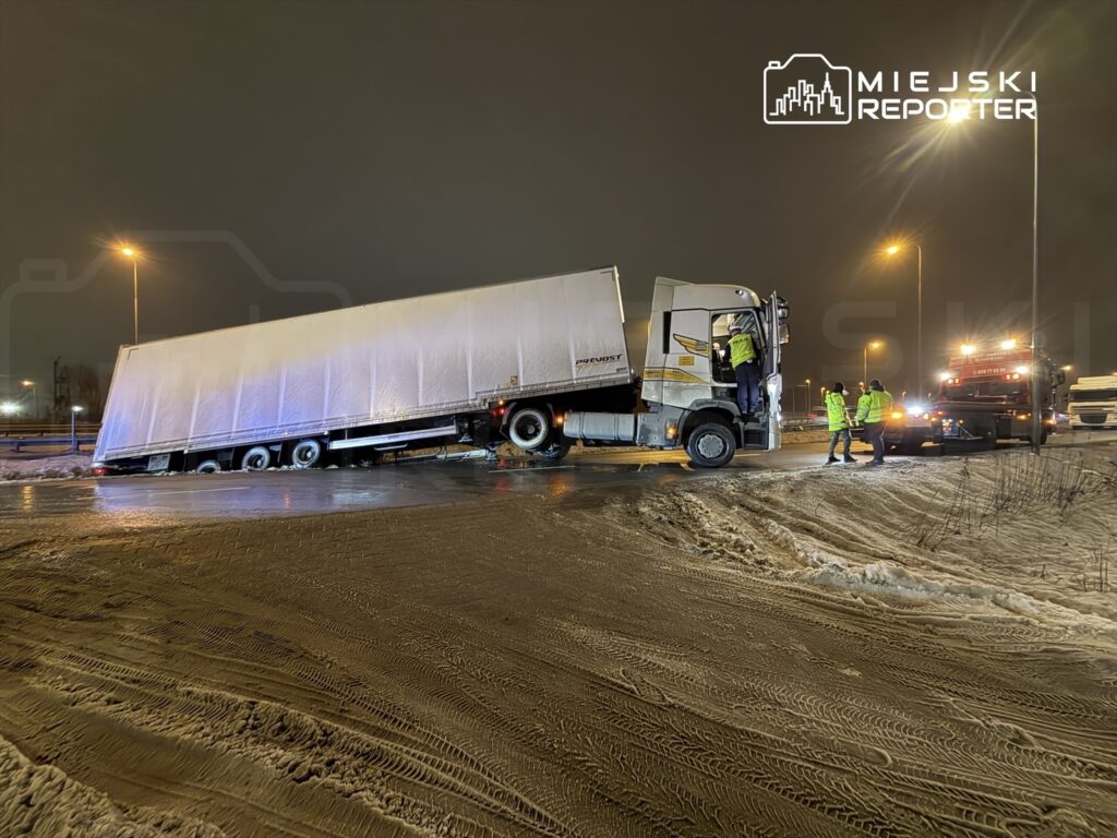 Ciągnik siodłowy z naczepą stoi na poboczu drogi, a grupa pracowników w odblaskowych kamizelkach zabezpiecza miejsce zdarzenia w nocy.