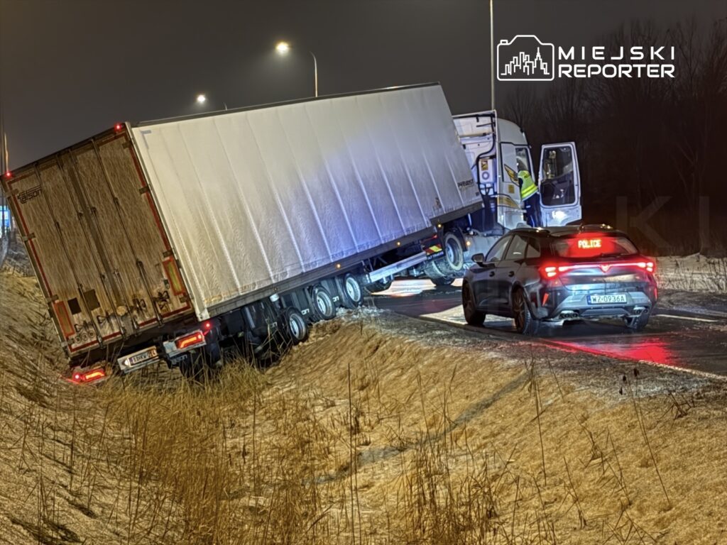 Ciężarówka z naczepą utknęła na poboczu drogi, obok niej stoi radiowóz policyjny z włączonymi światłami.
