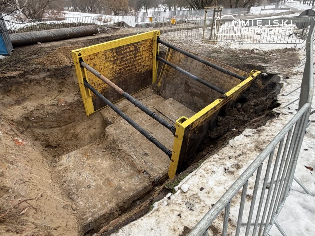 Wykop z żółtą osłoną zabezpieczającą, w tle widoczne śnieg i ogrodzenie budowlane.