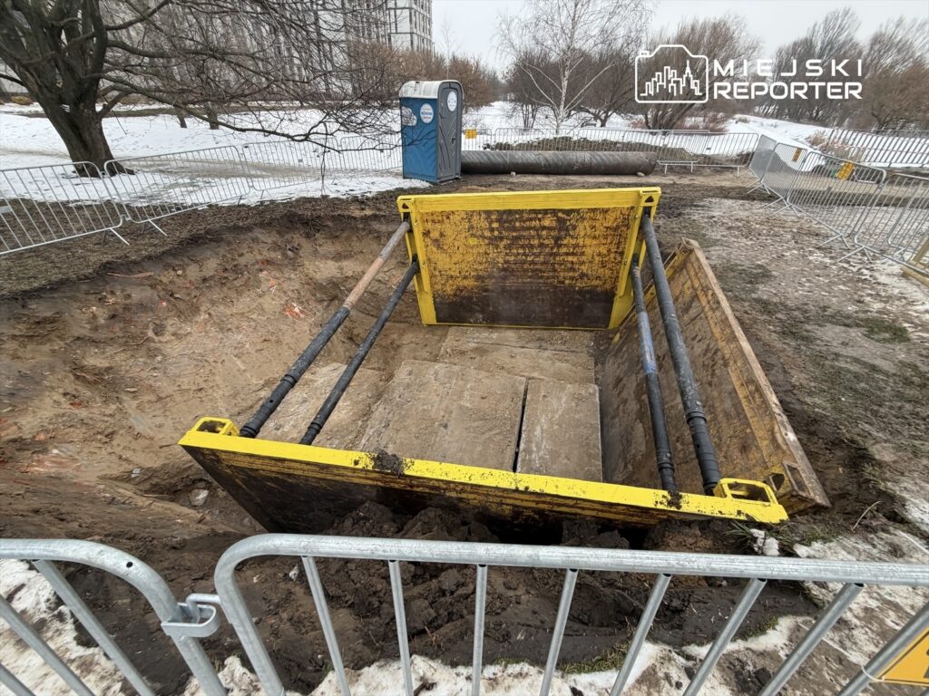 Wykop z żółtą skrzynią zabezpieczony barierkami, w tle widoczne drzewa i toaleta przenośna.