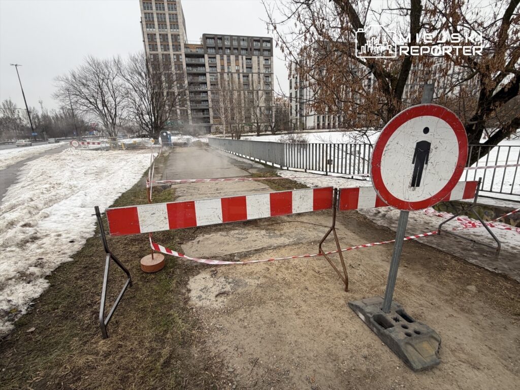 Zamknięta ścieżka piesza z barierkami i znakiem informacyjnym w otoczeniu budynków mieszkalnych i śniegu.