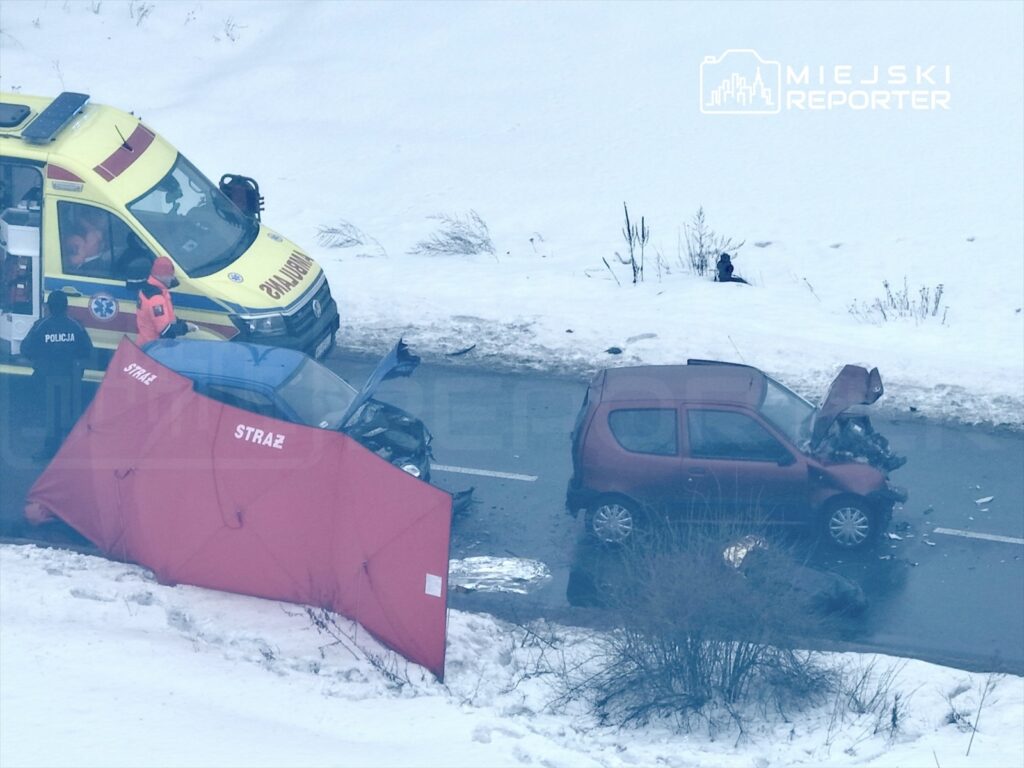 Zespół ratunkowy udziela pomocy po wypadku drogowym na zaśnieżonej drodze, obok uszkodzonego samochodu.