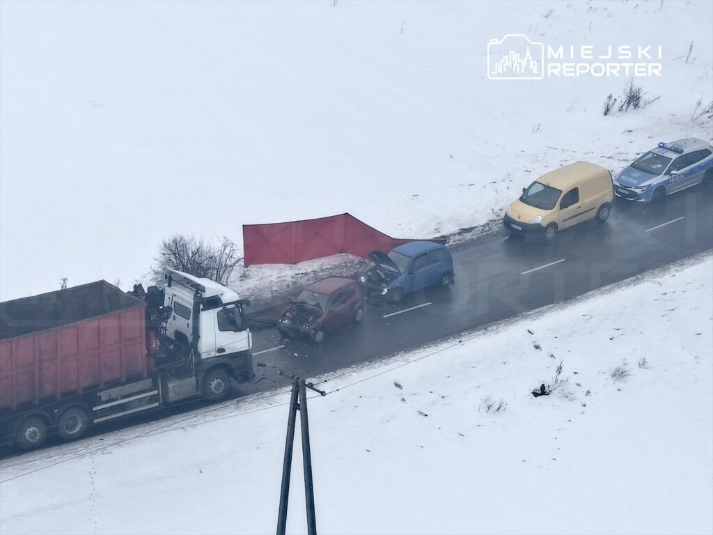 Wielka ciężarówka z czerwoną skrzynią stoi obok zderzonego samochodu osobowego na zaśnieżonej drodze. Obok pojazdu policyjnego znajduje się żółty furgon.