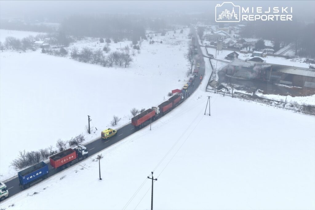 Długi sznur ciężarówek na zaśnieżonej drodze, obok nich pojazd służb ratunkowych, w tle rozmyty krajobraz.