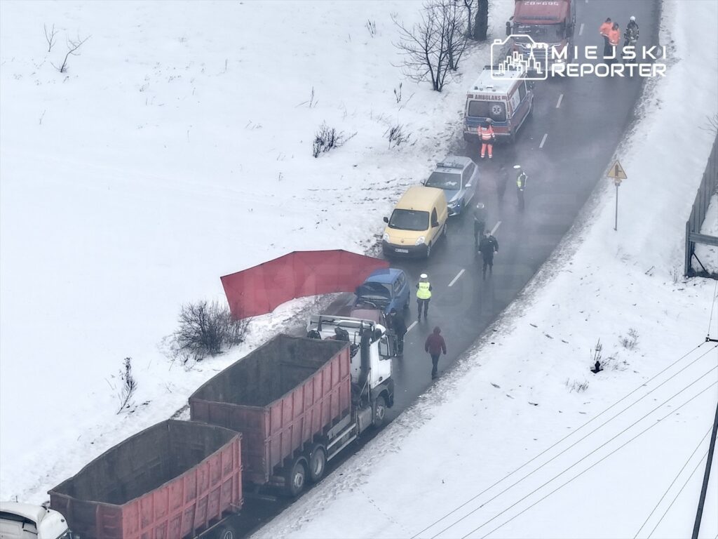 Grupa ludzi w odblaskowych kamizelkach rozmawia obok zablokowanej drogi, na której stoją samochody w śnieżnym otoczeniu.