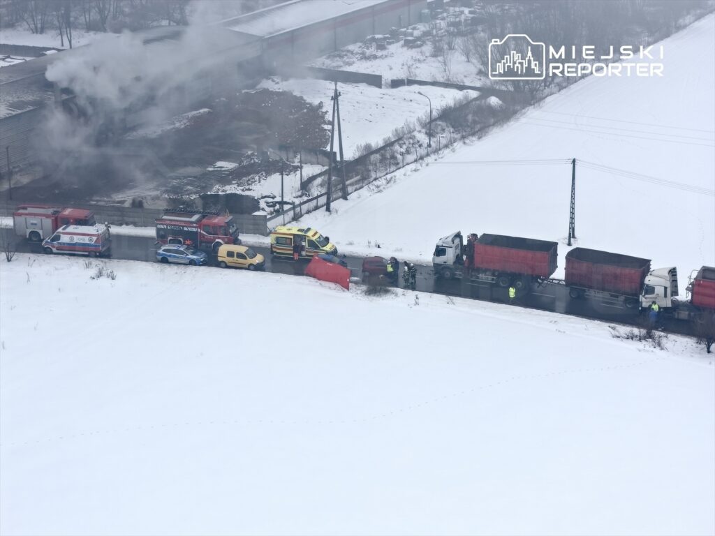 W akcji służby ratunkowe, w tym karetki i straż pożarna, przy drodze pokrytej śniegiem, obok przewróconego pojazdu.
