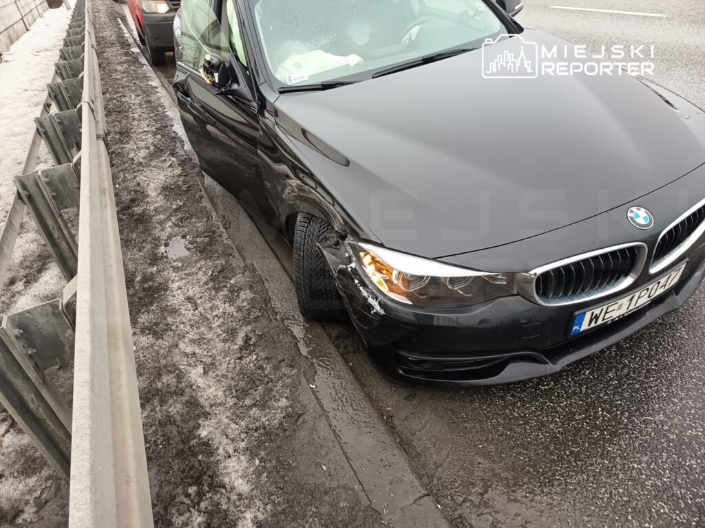 Czarny samochód osobowy zaparkowany blisko bariery ochronnej na mokrej jezdni, z widocznym uszkodzeniem nadkola.