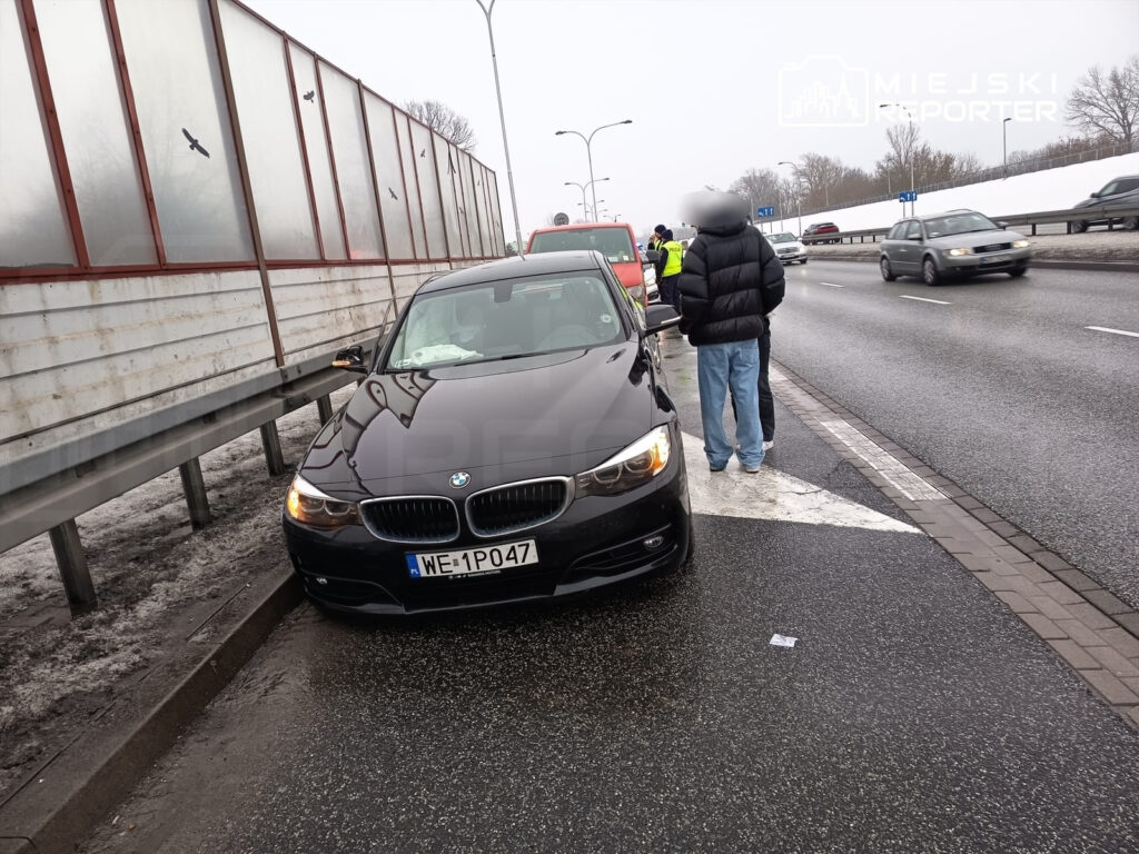 Czarny samochód osobowy zaparkowany na poboczu drogi, obok dwóch mężczyzn rozmawiających z policjantem.