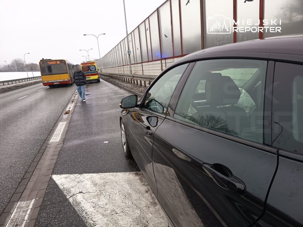 Mężczyzna w kurtce idzie wzdłuż drogi obok zatrzymanego autobusu i karetki, na mokrej nawierzchni.