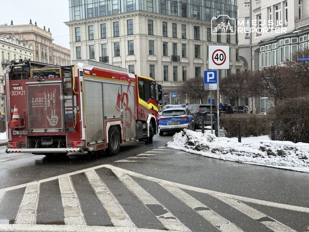 Straż pożarna z włączonymi sygnałami świetlnymi manewruje na zaśnieżonej ulicy, obok policyjnego radiowozu.