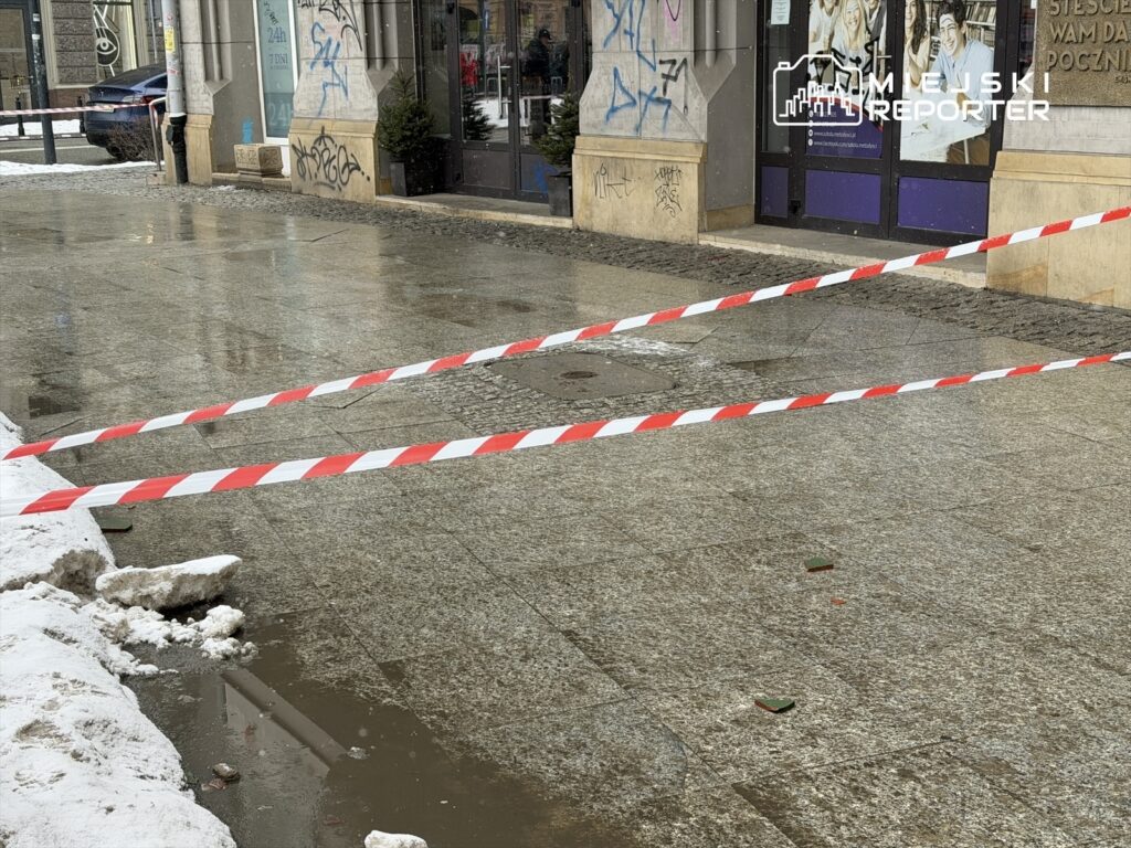 Na chodniku widać czerwoną i białą taśmę ostrzegawczą, oddzielającą zniszczoną nawierzchnię od reszty ulicy. W tle znajduje się budynek z graffiti oraz śnieg obok.