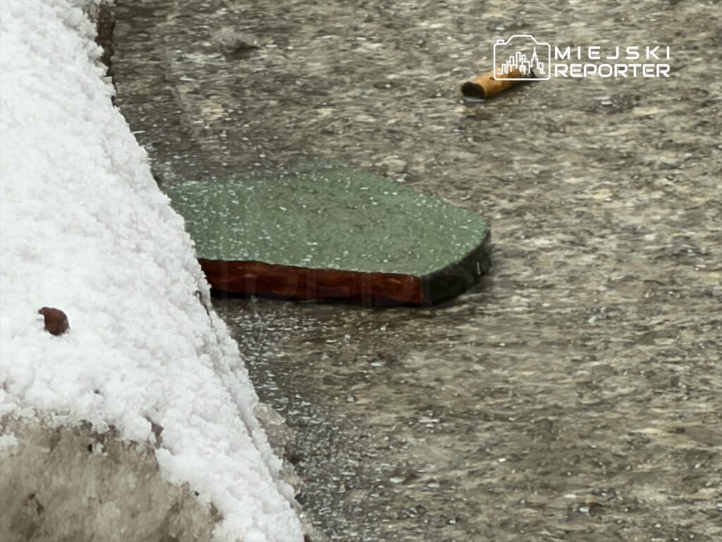 Na chodniku, obok śniegu, leży zielona płytka oraz niedopałek papierosa.
