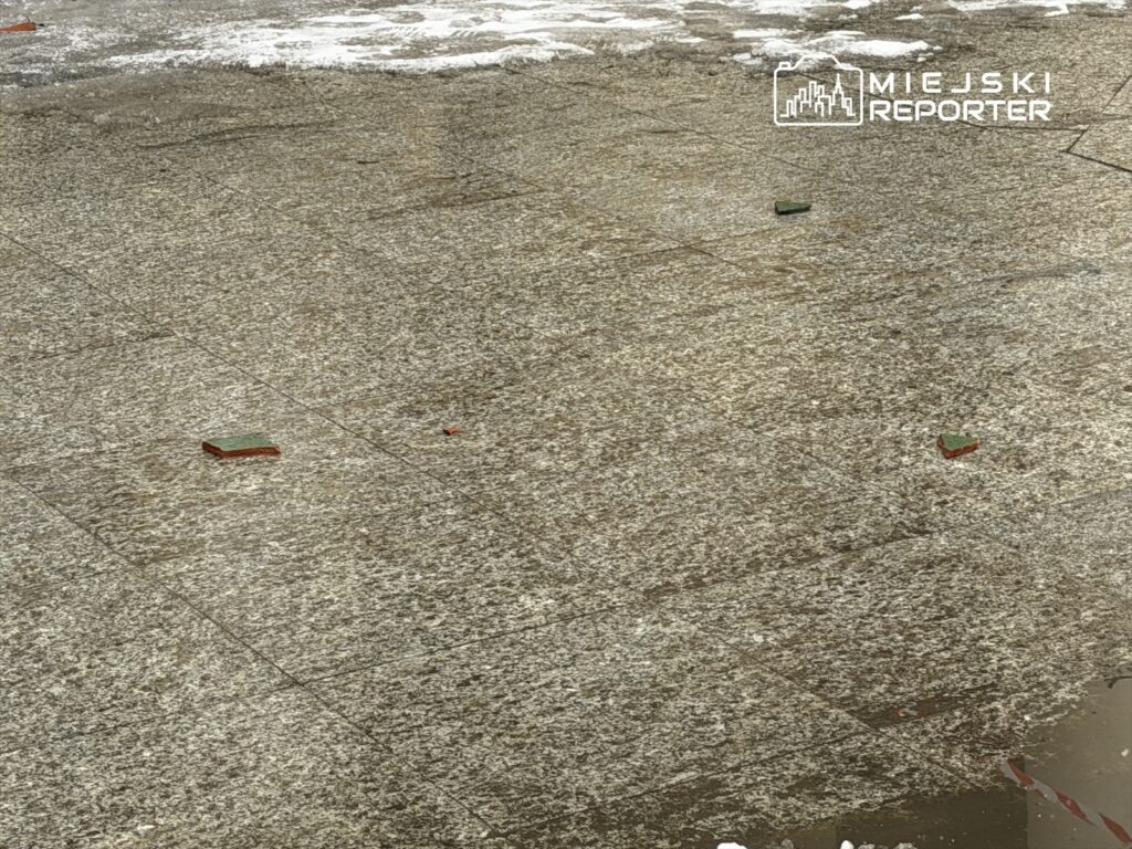 Na chodniku leżą fragmenty cegieł w kolorze zielonym i czerwonym, otoczone resztkami śniegu.