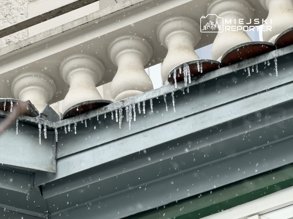 Sople lodu zwisające z krawędzi metalowego dachu, obok białe balustrady w tle, podczas deszczowej pogody.