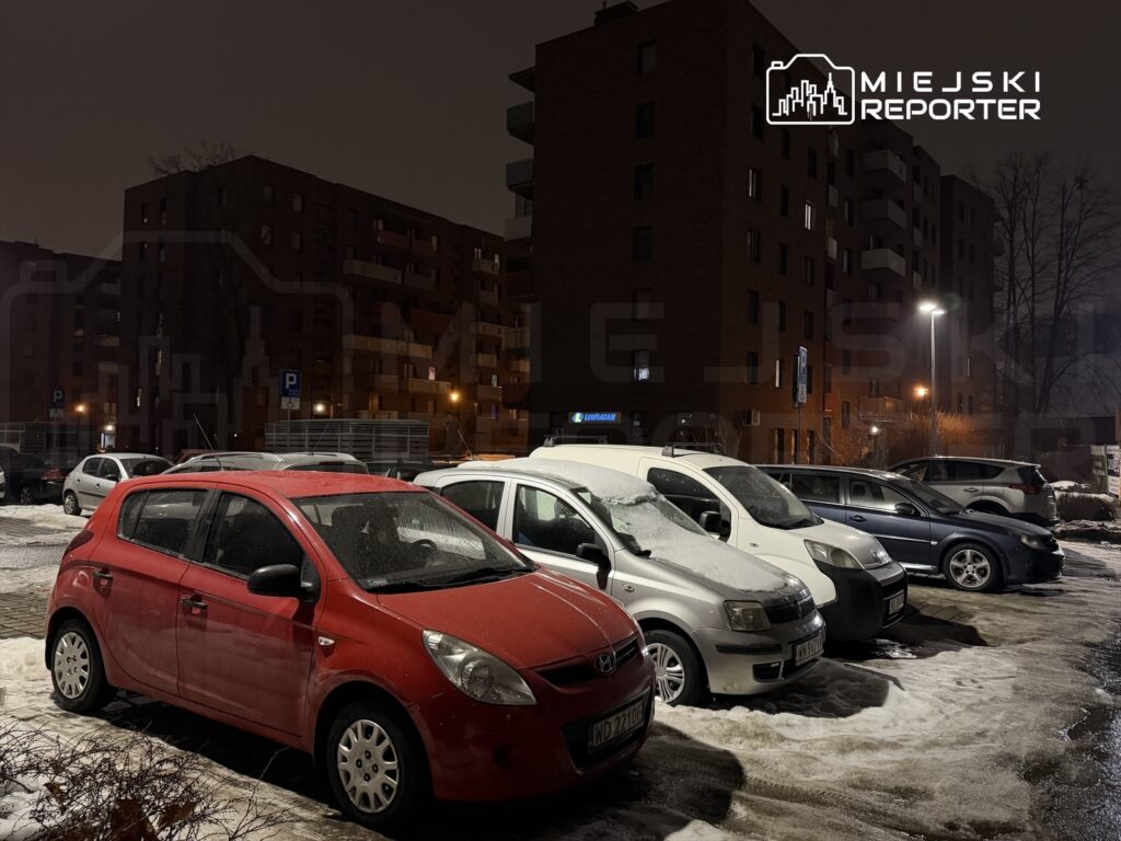 Parking z kilkoma samochodami, w tym czerwonym hatchbackiem, w oświetlonym nocą otoczeniu budynków mieszkalnych.