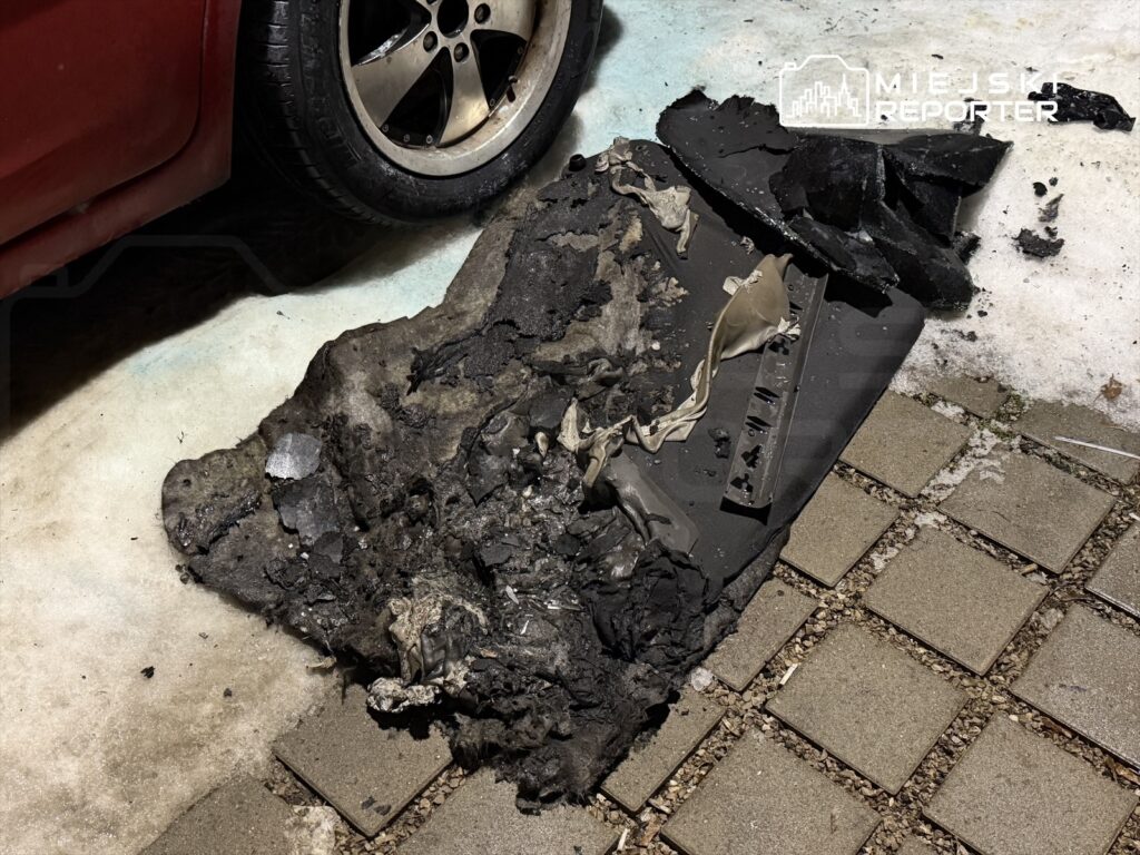 Uszkodzona opona leży na ziemi obok czerwonego samochodu, otoczona fragmentami czarnego materiału.