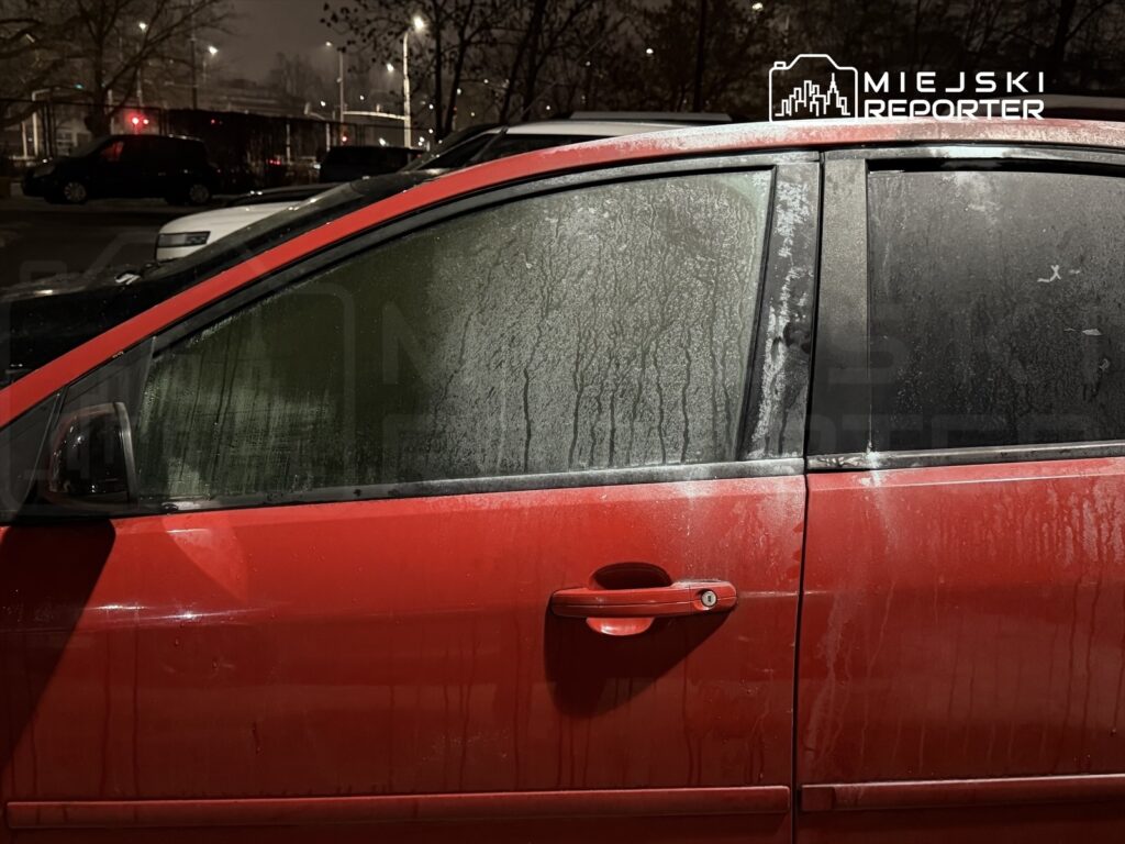 Czerwony samochód z zaparowanymi szybami zaparkowany na nocnej ulicy, w tle inne pojazdy.