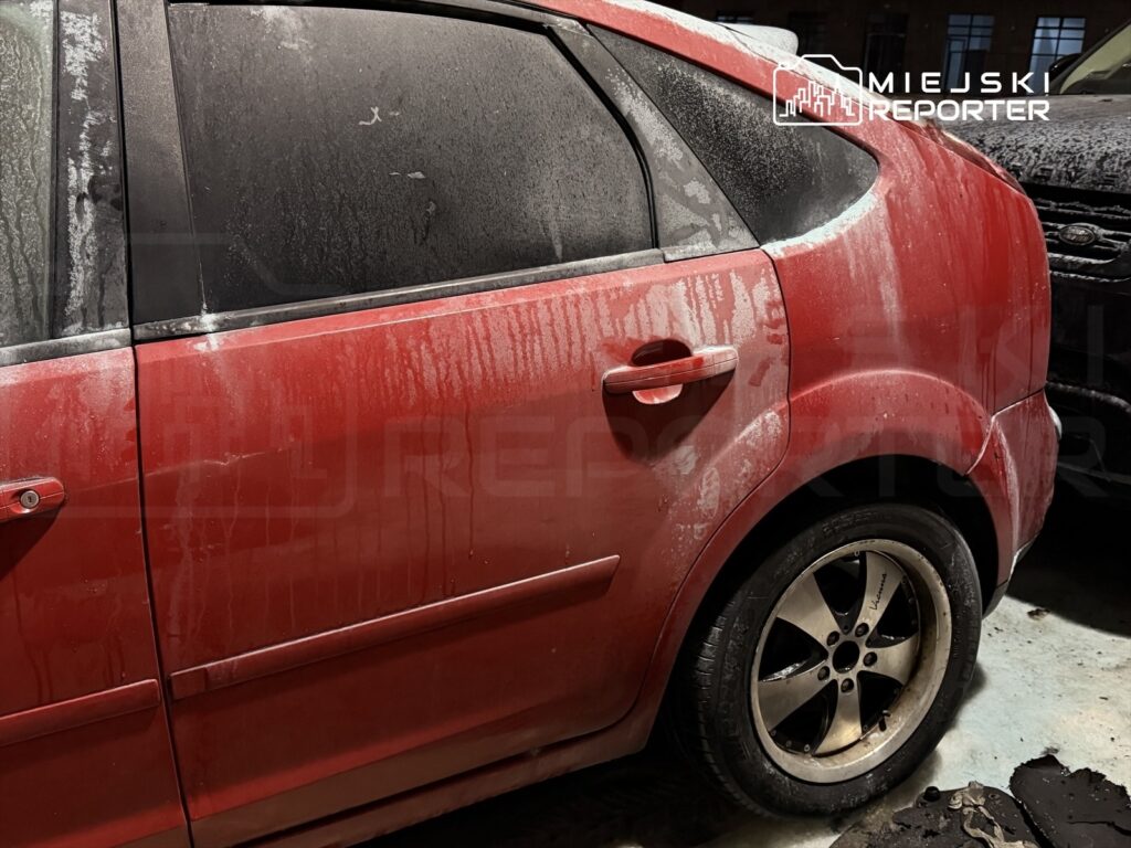 Czerwony samochód osobowy z zanieczyszczoną karoserią zaparkowany w ciemnym pomieszczeniu.