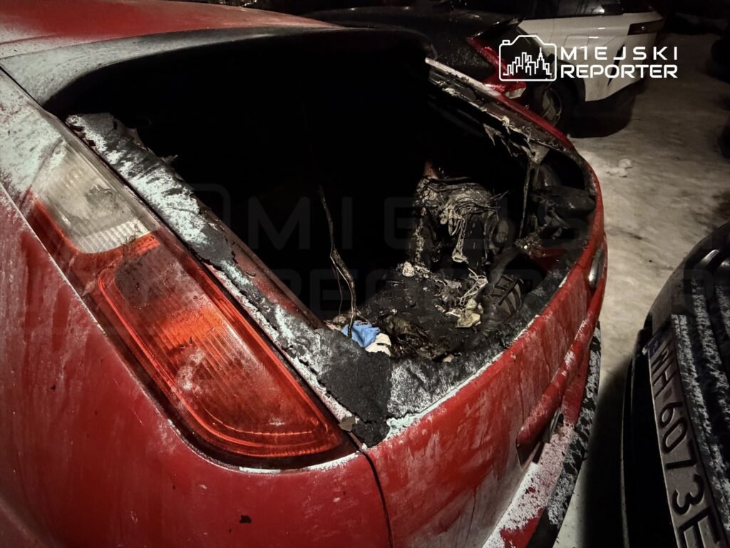 Uszkodzona tylna część czerwonego samochodu z widocznymi śladami po pożarze, w otoczeniu innych pojazdów na parkingu.