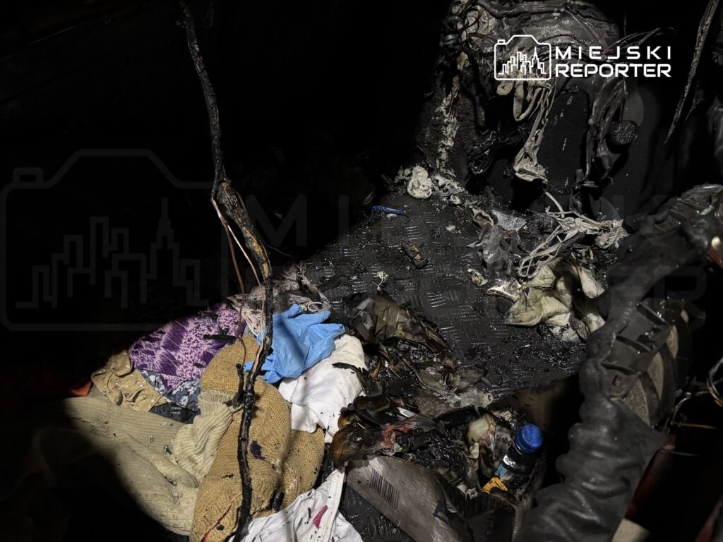 Zniszczona podłoga pojazdu z popalonymi materiałami, resztkami odzieży oraz uszkodzonymi przewodami.