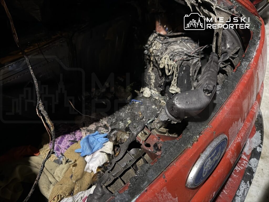 Zniszczony silnik czerwonego samochodu osobowego z widocznymi śladami pożaru i uszkodzonymi przewodami.