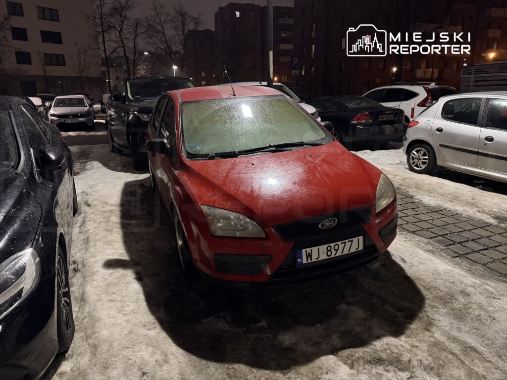 Czerwony samochód osobowy zaparkowany na ośnieżonym parkingu, w otoczeniu innych pojazdów.