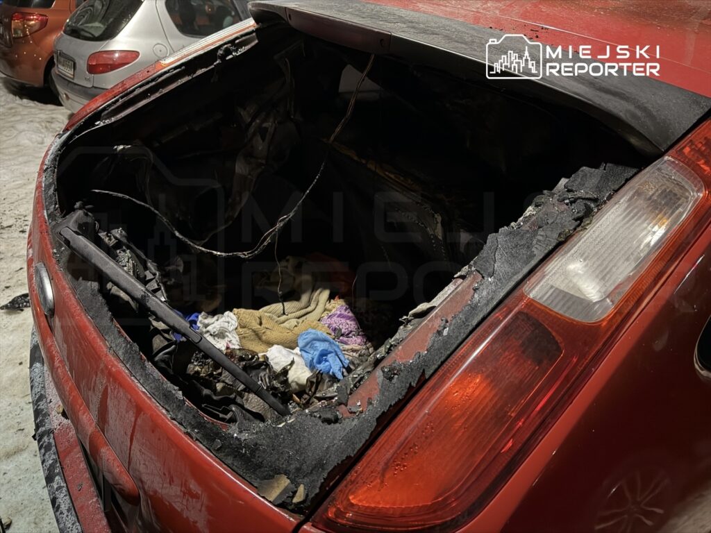 Czerwony samochód z uszkodzoną pokrywą bagażnika, w której widać spalone elementy i porozrzucane ubrania.