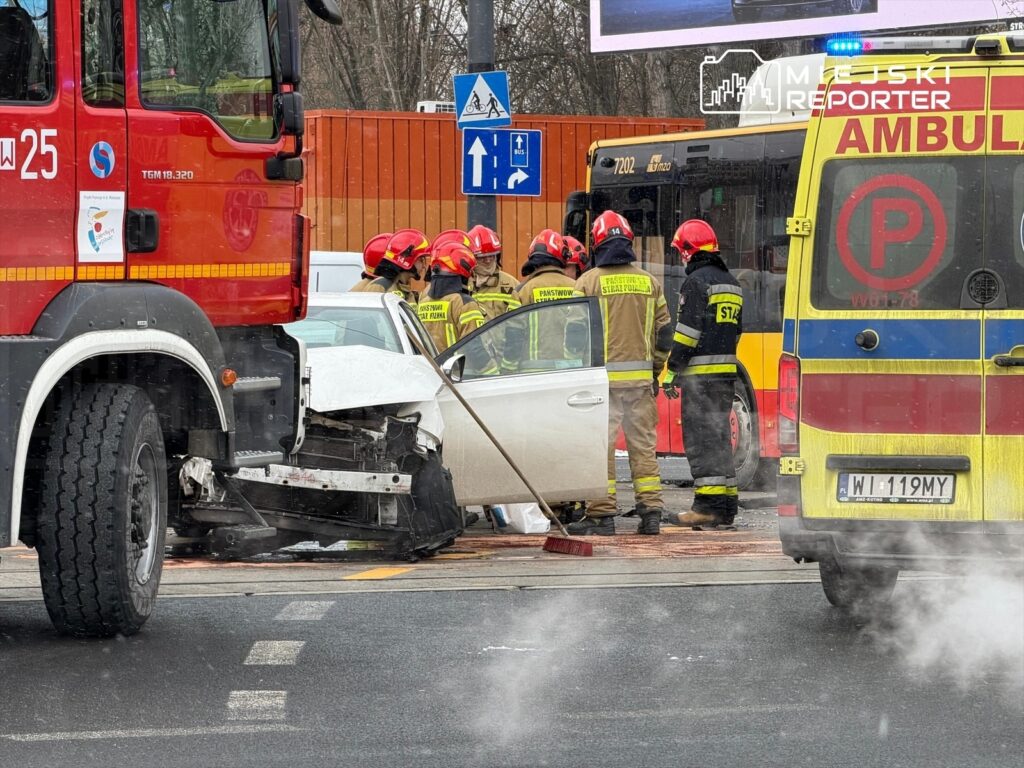 Grupa strażaków w kaskach i mundurach pracuje przy rozbitej białej limuzynie na skrzyżowaniu, obok stoi ambulans.