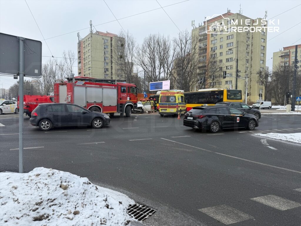 Na skrzyżowaniu zatrzymane pojazdy, w tym wóz strażacki i karetka, obok leży śnieg. W tle bloki mieszkalne.