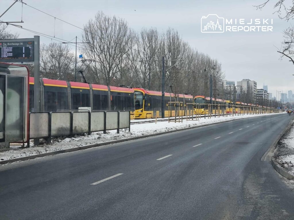 Tramwaje z czerwono-żółtymi dachami stoją na przystanku w miejskim otoczeniu, obok zaśnieżonej jezdni.