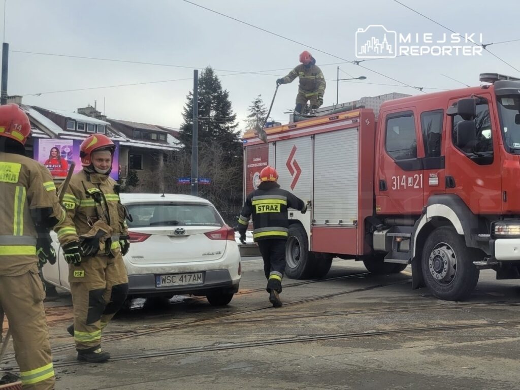 Grupa strażaków w mundurach, z hełmami, pracuje obok wozu strażackiego na miejskiej ulicy.
